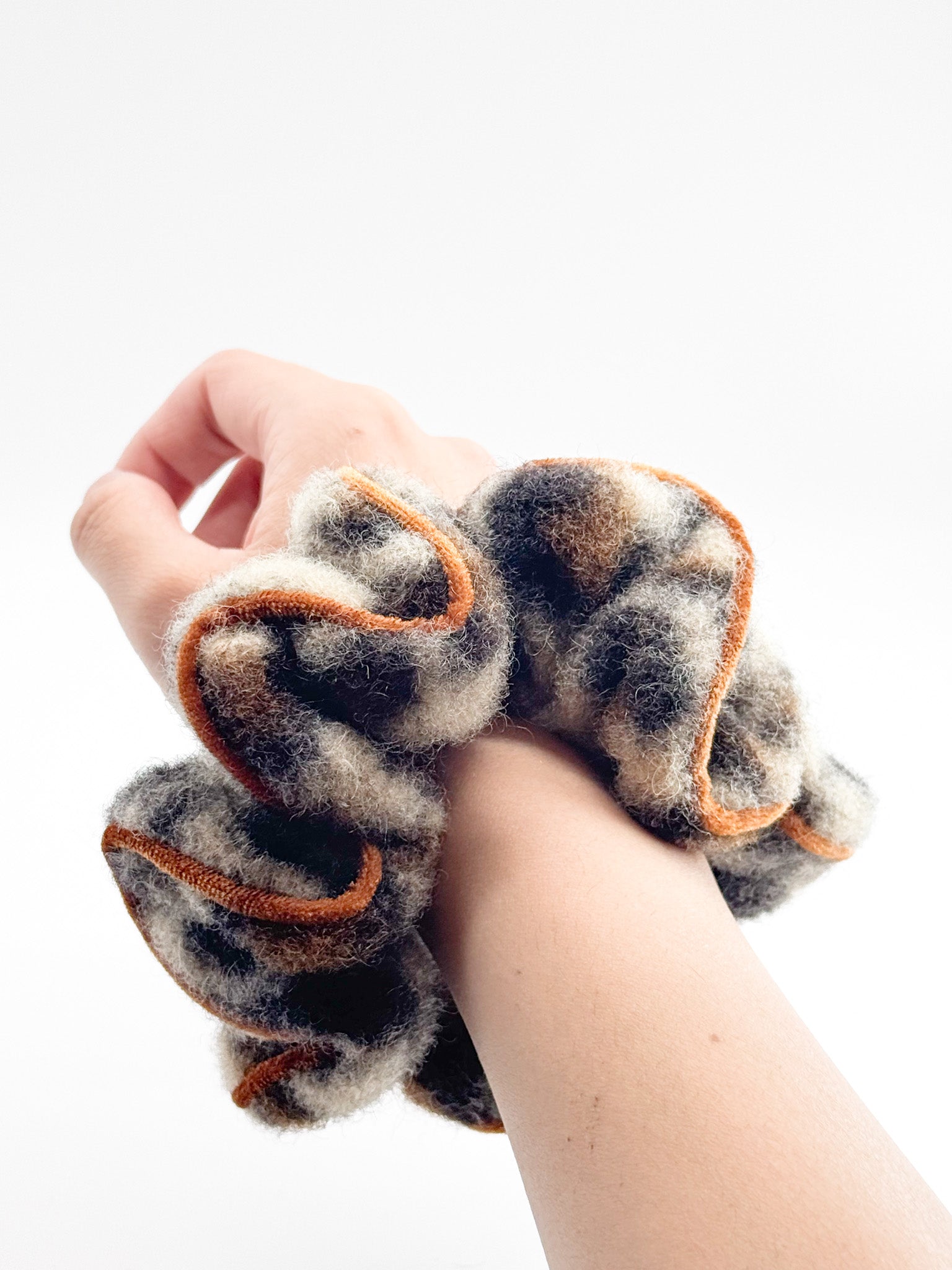 A leopard scrunchie with orange trim on a wrist against a white background